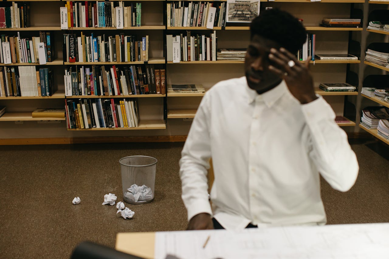 A stressed professional sits in a library workspace, surrounded by crumpled papers and bookshelves.