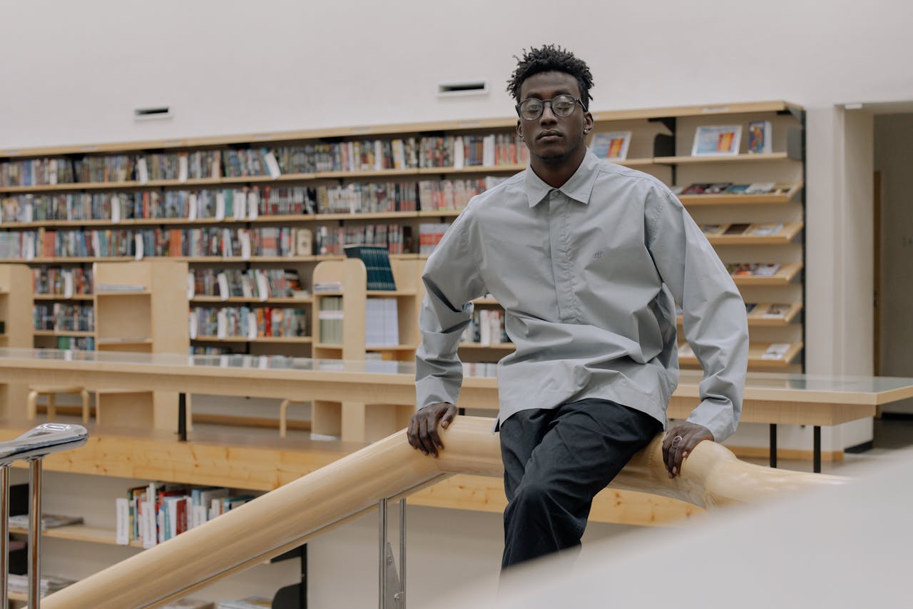 A young man sits confidently in a modern library, surrounded by bookshelves.